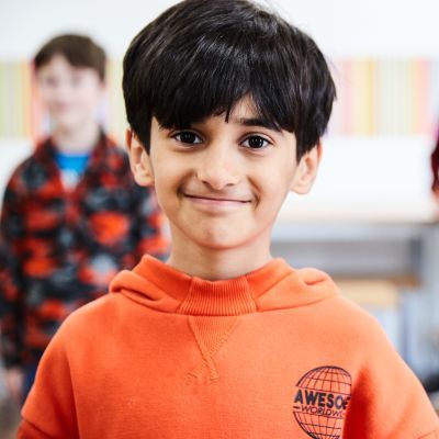 A young boy with black hair and wearing a bright orange jumper smiles at the camera
